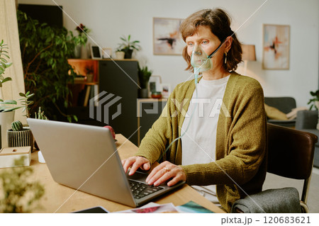 Senior woman using laptop at home while wearing oxygen mask, surrounded by cozy furniture and plants 120683621