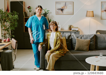 Portrait of elderly woman sitting on sofa with nurse standing beside her in a cozy living room setting featuring a lamp, paintings, and potted plants 120683680