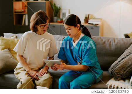 Nurse sitting on couch with elderly woman, discussing something on digital tablet in living room setting 120683780