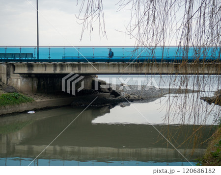 A man walks along the bridge along the seashore. Walking in the resort in the winter season. The man in the hood. Coast of the Black Sea. 120686182