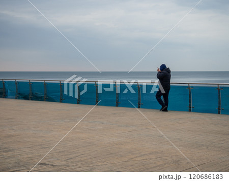A man walks along the bridge along the seashore. Walking in the resort in the winter season.  man in the hood speaks on the phone. Coast of the Black Sea. 120686183
