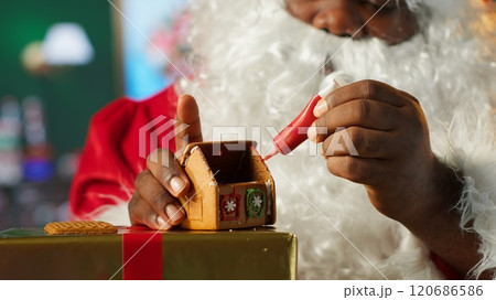Father Christmas decorates homemade gingerbread house with frosting and beads, North Pole cabin workshop. Black Santa keeping the festive tradition of baking sweet treats with cinnamon. Camera A. Father Christmas decorates homemade gingerbread house with frosting and beads, North Pole cabin workshop. Black Santa keeping the festive tradition of baking sweet treats with cinnamon. Camera A. 120686586