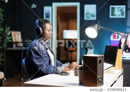 African American woman working on laptop, programming code while listening to music on wireless headset. Female freelancer with headphones using personal computer for monitoring system software. African American woman working on laptop, programming code while listening to music on wireless headset. Female freelancer with headphones using personal computer for monitoring system software. 120686621