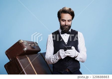 Hotel staff poses in studio with trolley baggage while he is cruising through social media and messaging. Doorkeeper or bellhop in official attire using phone app for surfing web pages. Hotel staff poses in studio with trolley baggage while he is cruising through social media and messaging. Doorkeeper or bellhop in official attire using phone app for surfing web pages. 120686663