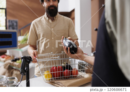 A bearded Caucasian man hands an organic aubergine to an employee for measuring. A male customer checks the weight of freshly harvested fruits and vegetables at the cashier's counter. A bearded Caucasian man hands an organic aubergine to an employee for measuring. A male customer checks the weight of freshly harvested fruits and vegetables at the cashier's counter. 120686671