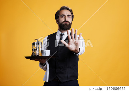 Skilled butler does stop sign with palm on camera, expressing disapproval and dismissing something. Young man with waiter occupation shows rejection and disagreement, not accept. Skilled butler does stop sign with palm on camera, expressing disapproval and dismissing something. Young man with waiter occupation shows rejection and disagreement, not accept. 120686683