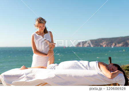 Therapist massaging woman's leg on table by sea 120686926