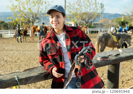 Portrait of positive young female stable worker holding horse halter or harness near wooden fence on ranch Portrait of positive young female stable worker holding horse halter or harness near wooden fence on ranch 120691948