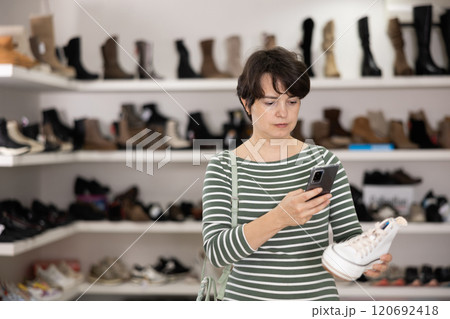 Female customer examines shoes in store and scans label with phone camera. Female customer examines shoes in store and scans label with phone camera. 120692418