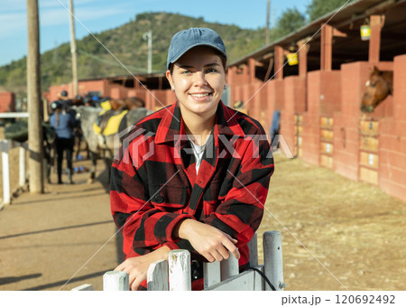 Portrait of positive smiling European young woman wearing plaid shirt and posing on yard of horse stables Portrait of positive smiling European young woman wearing plaid shirt and posing on yard of horse stables 120692492