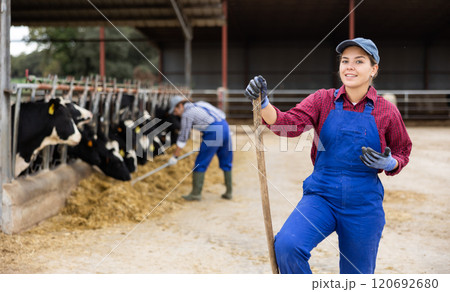 Portrait of smiling European female farmer in uniform with rake during work on dairy farm 120692680