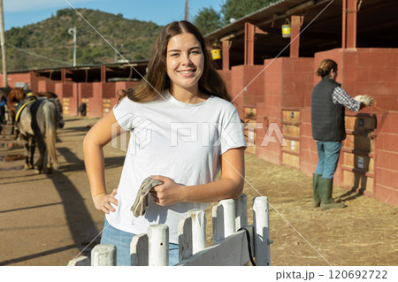 Happy young woman in casual clothes standing near fence and smiling at camera during visit of horse ranch Happy young woman in casual clothes standing near fence and smiling at camera during visit of horse ranch 120692722