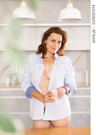 Topless woman in underpants and shirt stands in kitchen and covers bare chest with cloth of jacket. 120692834