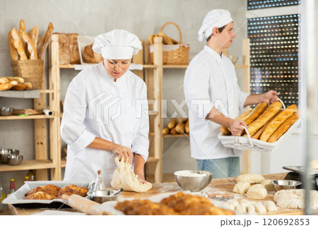 Female baker kneads dough on kitchen counter while young male assistant carries fresh baguettes to bakery display case 120692853
