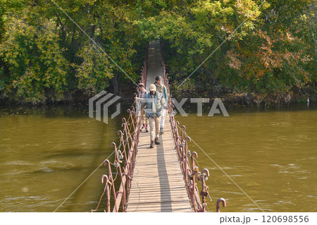 High angle shot of African American family with camping equipment going on wooden bridge over wide river in forest High angle shot of African American family with camping equipment going on wooden bridge over wide river in forest 120698556