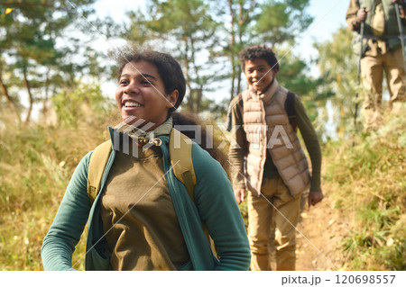 Medium close up of Mixed race n teen girl equipped with hiking backpack smiling widely while her younger brother and father following her Medium close up of Mixed race n teen girl equipped with hiking backpack smiling widely while her younger brother and father following her 120698557