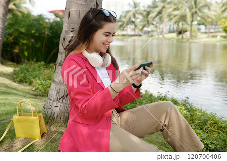 Smiling Young Woman Relaxing by Lake Holding Smartphone Smiling Young Woman Relaxing by Lake Holding Smartphone 120700406