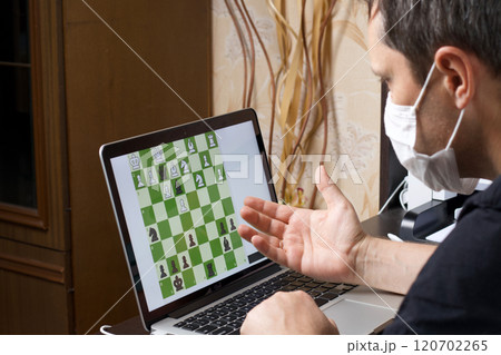 A man in a medical mask solves a chess problem online. Chess board with figures on a laptop screen. Leisure in self-isolation. International chess day. 120702265