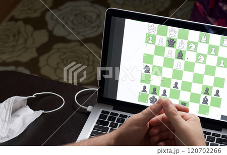A man solves a chess problem online. Chess board with figures on a laptop screen. Leisure in self-isolation, in the twilight. International chess day. A man solves a chess problem online. Chess board with figures on a laptop screen. Leisure in self-isolation, in the twilight. International chess day. 120702266