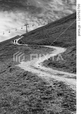 A winding alpine road stretches uphill, lined by grass and a cable car system above. A few hikers make their way along the path under a cloudy sky, enjoying the serene landscape. A winding alpine road stretches uphill, lined by grass and a cable car system above. A few hikers make their way along the path under a cloudy sky, enjoying the serene landscape. 120702700