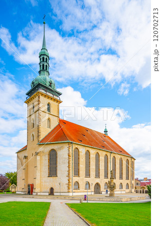 The Church of the Assumption of the Virgin Mary stands majestically in Most, showcasing late Gothic architecture under a bright blue sky and surrounded by lush greenery. 120702713