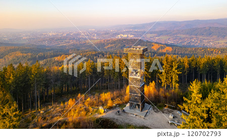 The wooden lookout tower stands tall against the vibrant autumn sunset near Liberec, surrounded by forested hills and distant views of the landscape bathed in warm colors. 120702793
