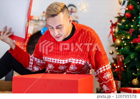 Christmas day concept. handsome man sitting surprised and opening gift box with christmas tree background. Festive atmosphere party. Christmas day concept. handsome man sitting surprised and opening gift box with christmas tree background. Festive atmosphere party. 120705578