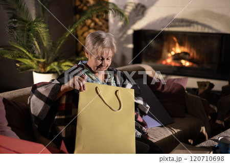 Elderly woman opening a yellow gift bag while sitting in a cozy living room near a glowing fireplace. Warm and festive atmosphere with a relaxed vibe. Elderly woman opening a yellow gift bag while sitting in a cozy living room near a glowing fireplace. Warm and festive atmosphere with a relaxed vibe. 120710698