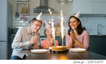 Family in festive hats claps and cheers while admiring the sparkling ice fountains on a birthday cake at home 120712077
