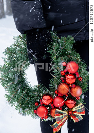 Unrecognizable woman holding Handmade Christmas wreath from red decorations. Festive holiday preparation outdoors in winter snowy weather. Sustainable responsible holiday gift 120713672
