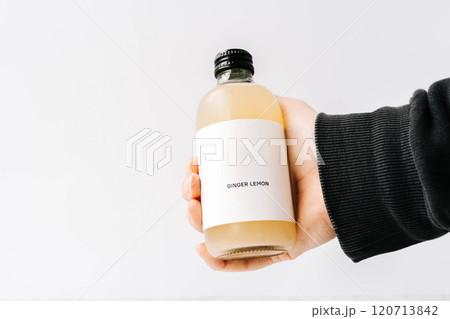 Woman hand holding small transparent glass bottle with black cap and white label, filled kombucha of yellow 120713842