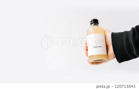 Woman hand holding small transparent glass bottle with black cap and white label, filled kombucha of yellow 120713843