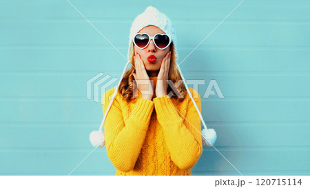 Winter portrait young woman blowing kiss wearing yellow knitted sweater and white hat on blue wall 120715114