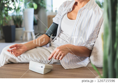 A middle-aged woman sits on the couch and measures her own blood pressure. Concept hypertension 120715596