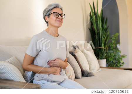 Mature beautiful middle-aged woman with gray hair sits on the couch and holds her stomach Mature beautiful middle-aged woman with gray hair sits on the couch and holds her stomach 120715632