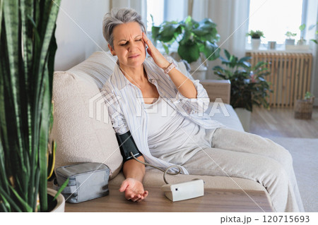 An elderly woman with gray hair sits on the couch and measures blood pressure with a tonometer. 120715693