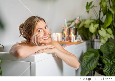 A young woman holds a glass of champagne while lying in a bath with foam and rose petals.  120717677