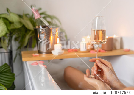 A young woman holds a glass of champagne while lying in a bath with foam and rose petals.  120717678