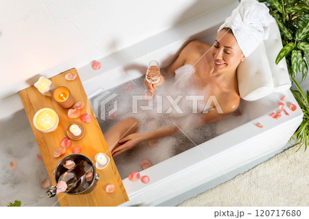 A young beautiful woman lies in a bath with foam and rose petals, reads a book and drinks champagne. A young beautiful woman lies in a bath with foam and rose petals, reads a book and drinks champagne. 120717680