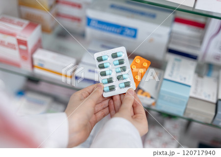 Hands of pharmacist holding medicine capsule blister pack at shelf stocked in pharmacy drugstore. Selecting type of medicine dispensing to patient. Hands of pharmacist holding medicine capsule blister pack at shelf stocked in pharmacy drugstore. Selecting type of medicine dispensing to patient. 120717940