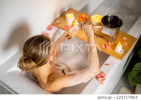 A young beautiful girl drinks wine while lying in a bath with foam and petals. Candles and plants 120717963