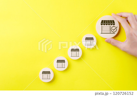 Business expansion and franchise growth concept. Hand selecting large store icon among smaller ones on yellow background, symbolizing franchise business growth, expansion strategy, market selection, Business expansion and franchise growth concept. Hand selecting large store icon among smaller ones on yellow background, symbolizing franchise business growth, expansion strategy, market selection, 120726152