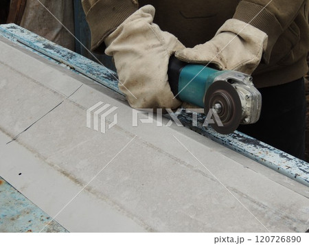 Hands in canvas gloves holding a grinder over a sheet of iron, sawing metal in an outdoor workshop in a village 120726890