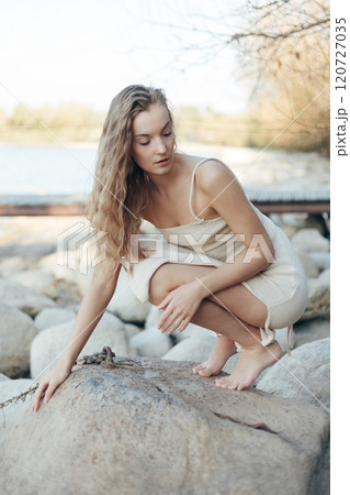 Young lonely sad blonde woman in knitted dress sitting on beach hugging legs resting chin on knees, with closed eyes 120727035