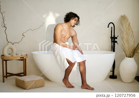 Young adult man with white towel on waist sitting on bath while checking water temperature in aesthetic bathroom 120727956