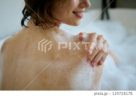Medium close up of unrecognizable smiling woman washing her body with aromatic shower gel while sitting in hot bathtub 120728176