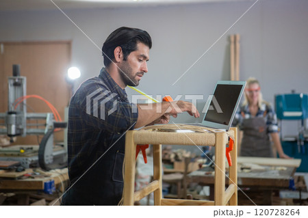 young carpenter caucasian man with laptop make drawing on wood to design at workspace. craftsman profession in wood factory 120728264