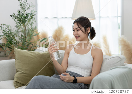 Asian young female sporty holding water bottle sitting on sofa in living room while exercising. stay hydrated per day. Healthy fit woman fresh wellness. Drinking water makes you feel good. Asian young female sporty holding water bottle sitting on sofa in living room while exercising. stay hydrated per day. Healthy fit woman fresh wellness. Drinking water makes you feel good. 120728787