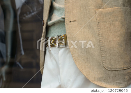 A mannequin dressed in trousers, a jacket, a belt and a jumper is standing behind glass in a shop window. 120729046