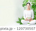 A serene woman in white meditating among lush greenery while holding a bowl of fresh herbs 120730507